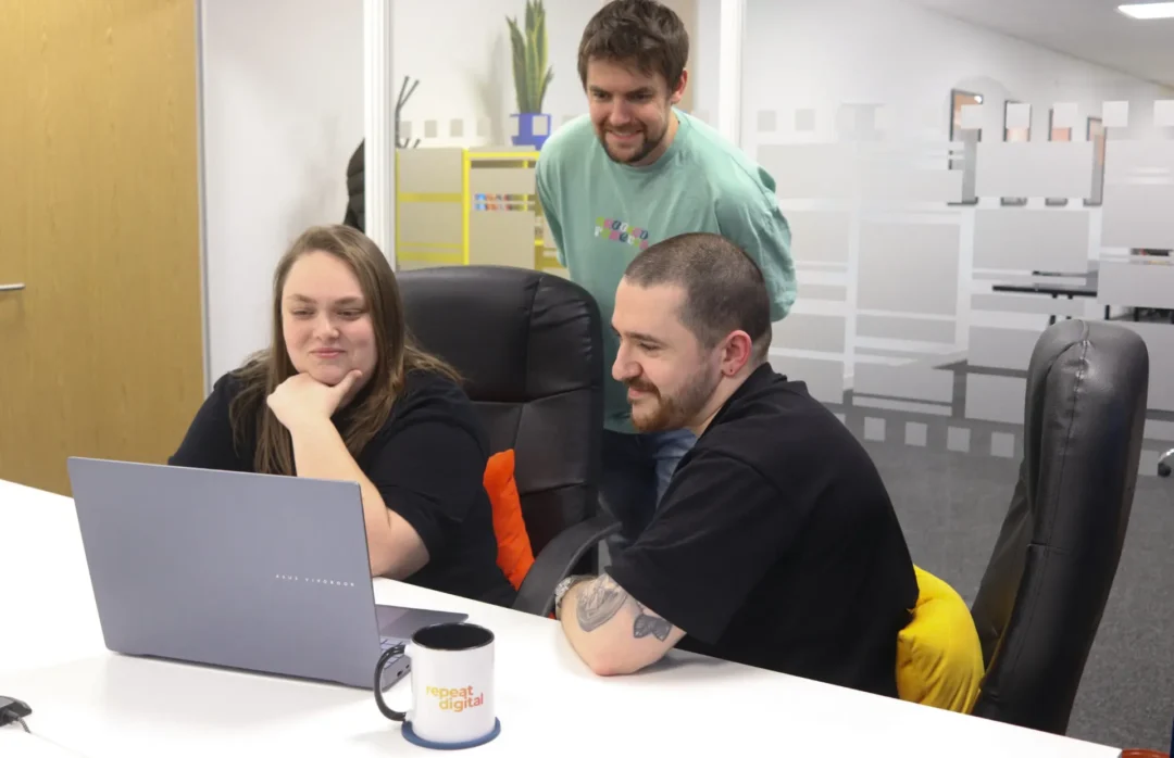 Three members of the Repeat Digital team collaborating and smiling while looking at a laptop in their Nottingham office.