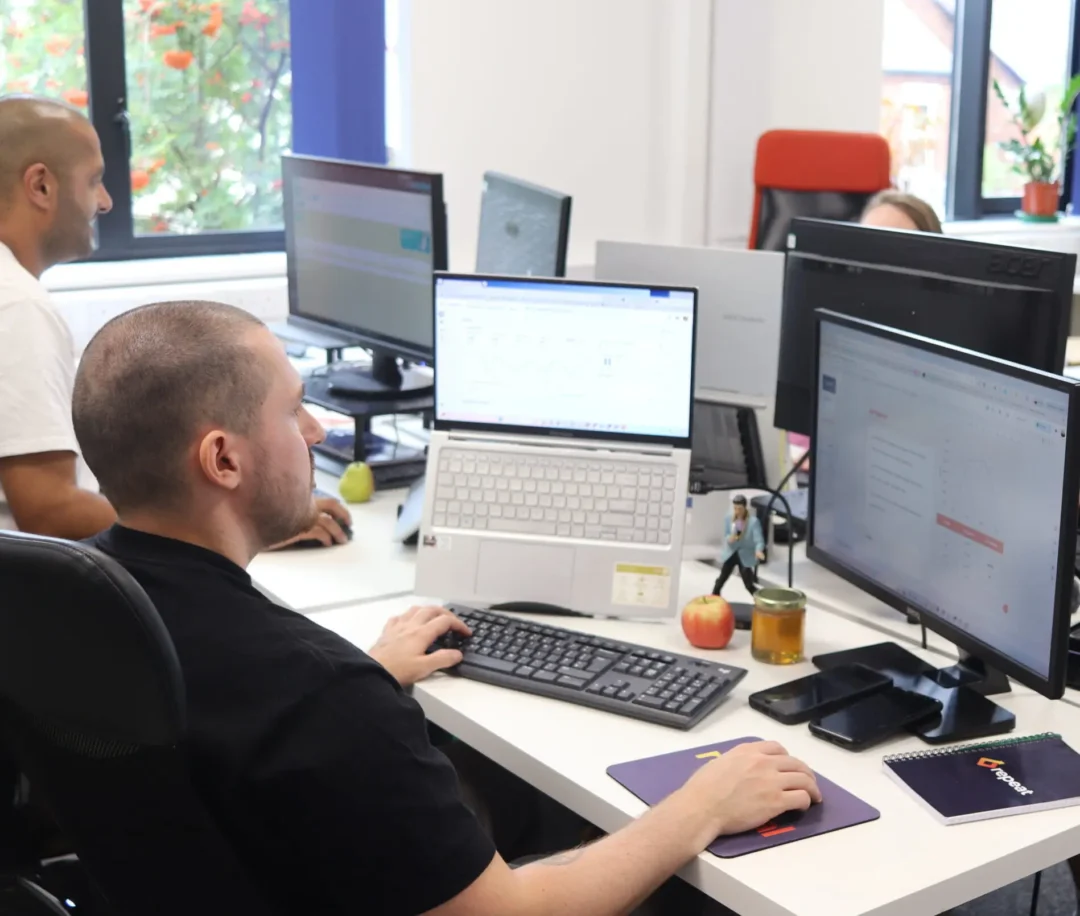 A digital marketing specialist at Repeat Digital working at a desk with a laptop and a large desktop monitor displaying website data and campaign performance.