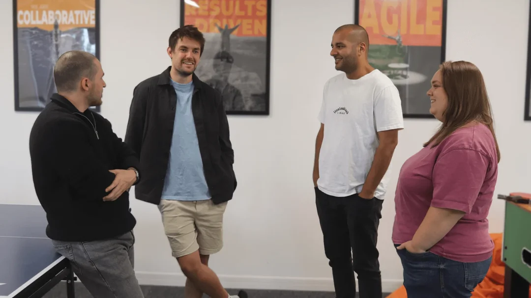 Four team members in a casual conversation in the office, standing in front of posters that read 'Collaborative', 'Results Driven', and 'Agile', representing the supportive working environment at Repeat Digital.